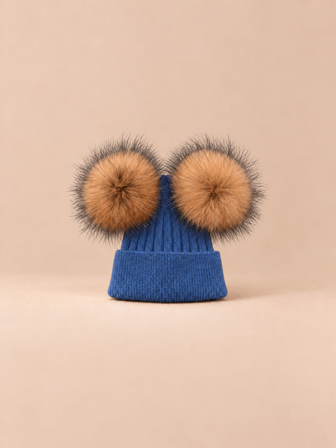 Bonnet double pompon, bleu, pompons naturels, vue de face sur fond pêche sable.