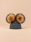 Bonnet double pompon, gris foncé, pompons naturels, vue de face sur fond pêche sable.